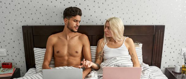 Blonde caucasian girl talking to thoughtful middle eastern man during they using laptops on bed at home. Young multiracial couple. Domestic entertainment and leisure. Moden spacious bedroom. Morning - Photo, Image