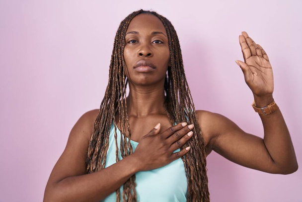 Африканська американка, що стоїть над рожевим фоном, лаючись руками на грудях і відкриваючи долоню, обіцяючи свою вірність.  - Фото, зображення