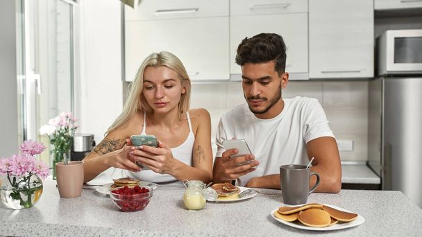 Young focused multiracial couple using smartphones during having breakfast at table at home kitchen. Middle eastern man and blonde caucasian girl. Domestic lifestyle. Modern apartment. Morning time - Photo, image