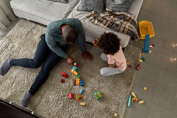 Top view of black father and son playing with construction building blocks on carpet at home. Man and child spending time together. Fatherhood and parenting. Domestic leisure, hobby and entertainment - Zdjęcie, obraz