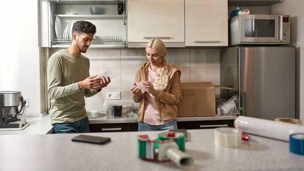 Young focused multiracial couple packing glasses in air bubble film for packing in cardboard box with name kitchen. Caucasian girl and middle eastern man moving to new apartment. Home relocation - Fotoğraf, Görsel