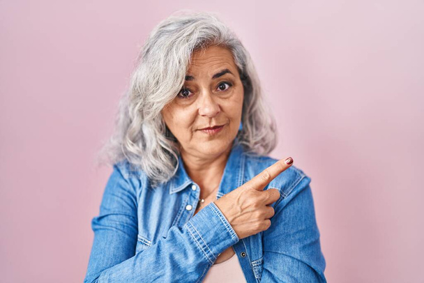 Mujer de mediana edad con el pelo gris de pie sobre fondo rosa apuntando con el dedo de la mano a un lado mostrando publicidad, cara seria y tranquila  - Foto, Imagen