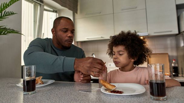 Adult father giving pizza slice to his little son during having lunch or dinner at table at home kitchen. Unhealthy eating. Young black family lifestyle and relationship. Fatherhood and parenting - Foto, Imagen