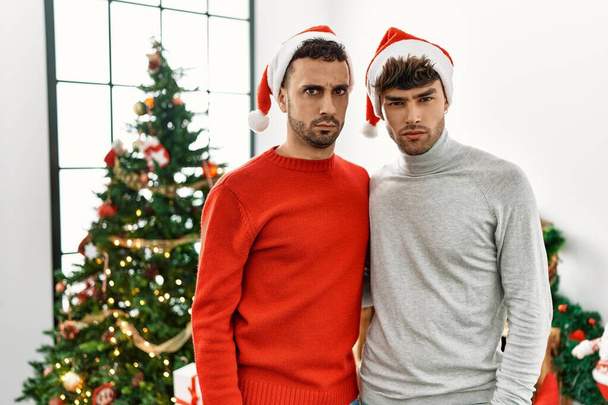 Young gay couple standing by christmas tree wearing hat skeptic and nervous, frowning upset because of problem. negative person.  - Photo, Image