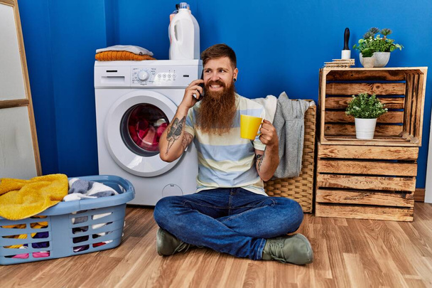 Молодий рудий чоловік розмовляє по смартфону і п'є каву в пральні
 - Фото, зображення