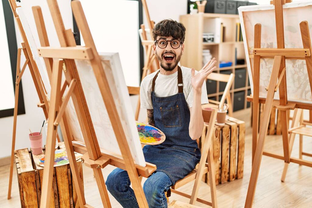 Spanischer Mann mit Bart im Kunststudio feiert Sieg mit glücklichem Lächeln und Siegerausdruck mit erhobenen Händen  - Foto, Bild