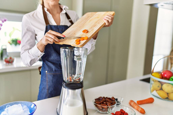 Young blonde woman pouring carrot on blender at kitchen - Photo, Image