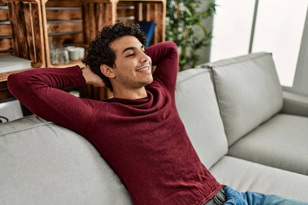 Νεαρός Ισπανόφωνος χαλαρώνει με τα χέρια στο κεφάλι κάθεται στον καναπέ στο σπίτι. - Φωτογραφία, εικόνα