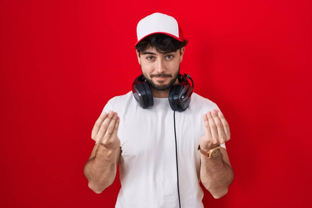 Hispanic man with beard wearing gamer hat and headphones doing money gesture with hands, asking for salary payment, millionaire business  - Photo, Image