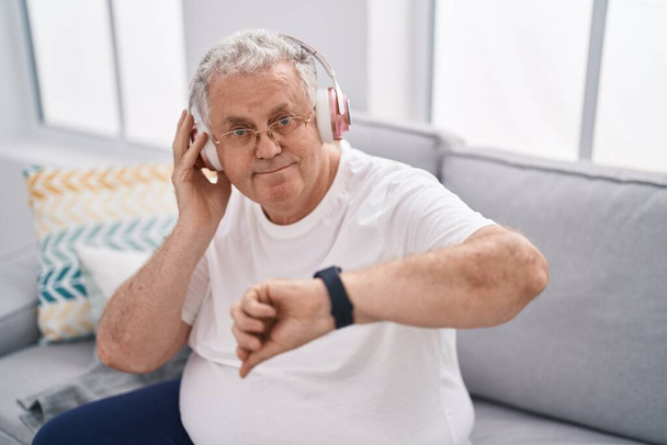 Middle age grey-haired man listening to music looking watch at home - Photo, Image
