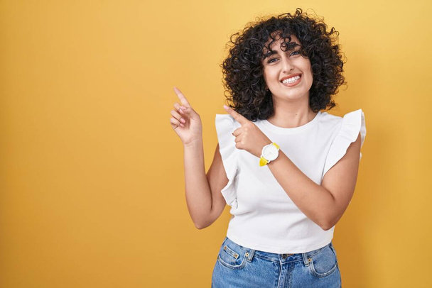 Young middle east woman standing over yellow background smiling and looking at the camera pointing with two hands and fingers to the side.  - Fotoğraf, Görsel