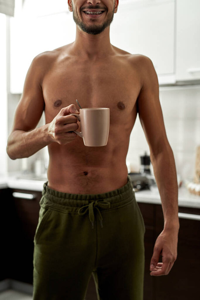 Obscure face of young smiling handsome man standing with cup of tea and coffee at home kitchen. Partial of bearded guy with naked torso. Domestic lifestyle. Spacious apartment. Daytime - Foto, immagini