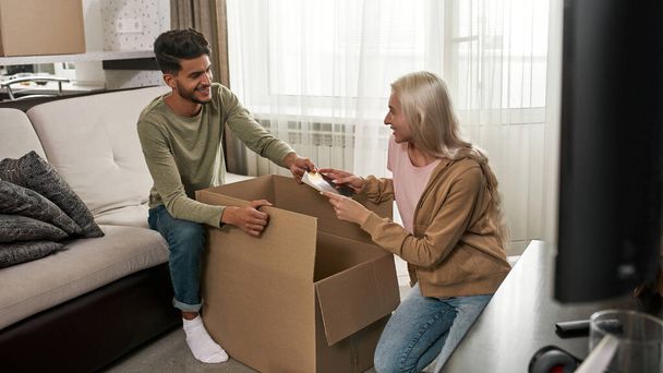 Middle eastern man taking book from caucasian girl for packing in cardboard box. Woman and guy looking at each other. Young smiling multiracial couple moving to new apartment. Home relocation - Fotoğraf, Görsel