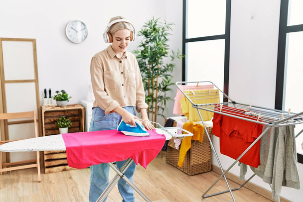 Young caucasian woman listening to music ironing clothes at laundry room - Foto, immagini