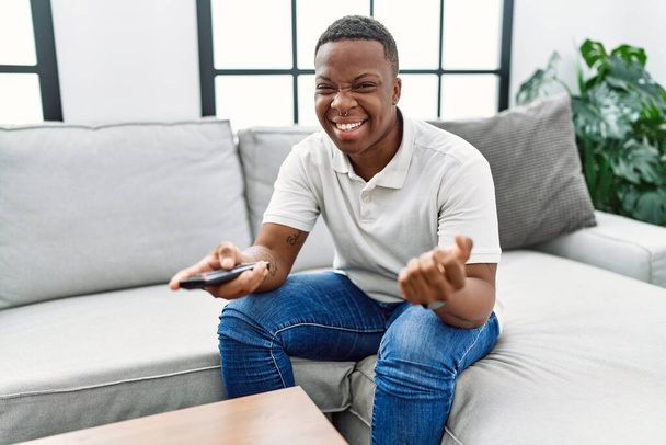 Jovem homem africano assistindo tv segurando controle remoto de televisão gritando orgulhoso, celebrando vitória e sucesso muito animado com braço levantado  - Foto, Imagem