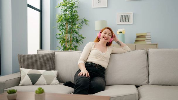 Νεαρή κοκκινομάλλα γυναίκα που ακούει μουσική καθισμένη στον καναπέ στο σπίτι - Φωτογραφία, εικόνα