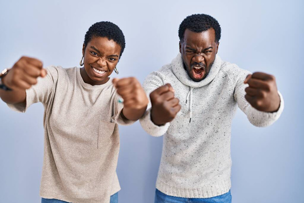 Νεαρό ζευγάρι Αφροαμερικανών στέκεται πάνω από το μπλε φόντο μαζί θυμωμένος και τρελός ανύψωση γροθιές απογοητευμένοι και έξαλλος, ενώ φωνάζει με θυμό. οργή και επιθετική αντίληψη.  - Φωτογραφία, εικόνα