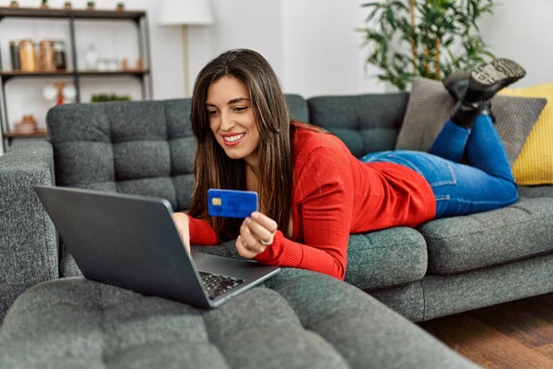 Mujer joven usando el ordenador portátil y la tarjeta de crédito tumbado en el sofá en casa - Foto, Imagen