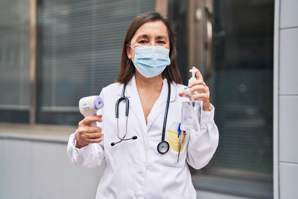 Жінка середнього віку в уніформі лікаря та медичній масці, що тримає термометр та дезінфікує гелеві руки на вулиці
 - Фото, зображення