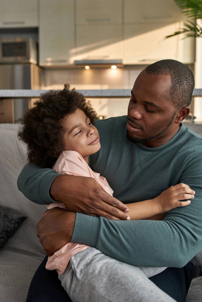Adult father embracing smiling little son on sofa at home. Black man and child looking at each other. Family closeness and tenderness. Fatherhood and parenting. Domestic leisure and entertainment - Photo, image