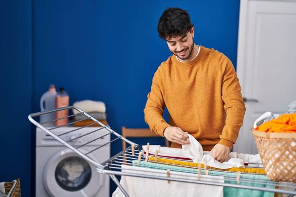 Молодий іспаномовний чоловік посміхається впевнено висить одяг на лінії одягу в пральні
 - Фото, зображення