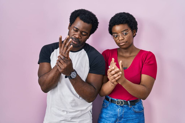 jong Afrikaans amerikaans paar staan over roze achtergrond lijden pijn op handen en vingers, artritis ontsteking  - Foto, afbeelding