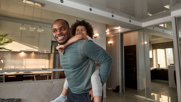 Adult father carrying his little son piggyback at home. Joyful black man and child playing, resting, joking and having fun together. Fatherhood and parenting. Domestic leisure and entertainment - Fotoğraf, Görsel