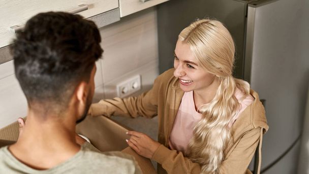 Young couple looking at each other during packing things in cardboard box at kitchen. Smiling pretty caucasian blonde girl and man moving to new apartment. Concept of home relocation - Foto, afbeelding