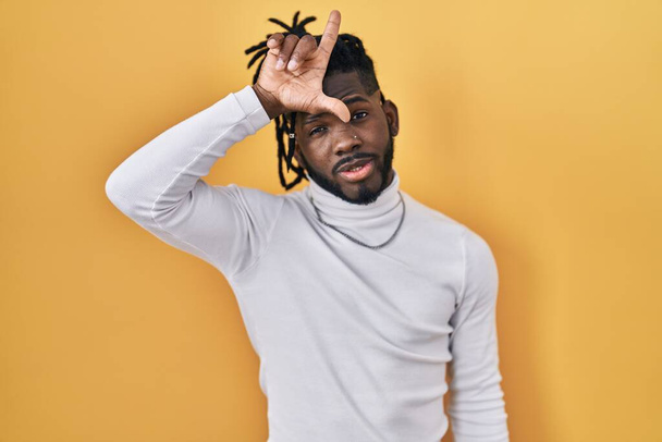 Afrikanischer Mann mit Dreadlocks trägt Rollkragenpullover über gelbem Hintergrund und macht sich über Menschen mit Fingern auf der Stirn lustig, die sich über Verlierer lustig machen und beleidigen.  - Foto, Bild