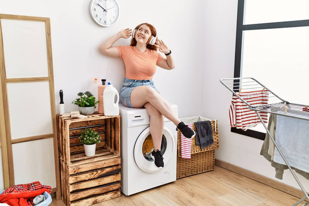 Junge rothaarige Frau hört Musik und wartet in Waschküche auf Waschmaschine - Foto, Bild