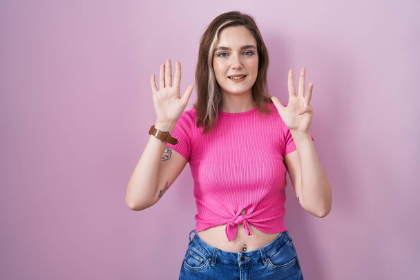 Blonde kaukasische Frau, die vor rosa Hintergrund steht und mit den Fingern Nummer neun nach oben zeigt, während sie selbstbewusst und glücklich lächelt.  - Foto, Bild
