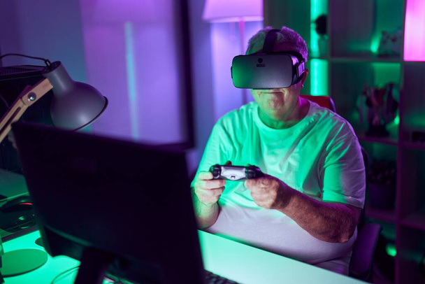 Homme aux cheveux gris moyen âge streamer jouer à un jeu vidéo en utilisant des lunettes de réalité virtuelle et joystick à la salle de jeux - Photo, image