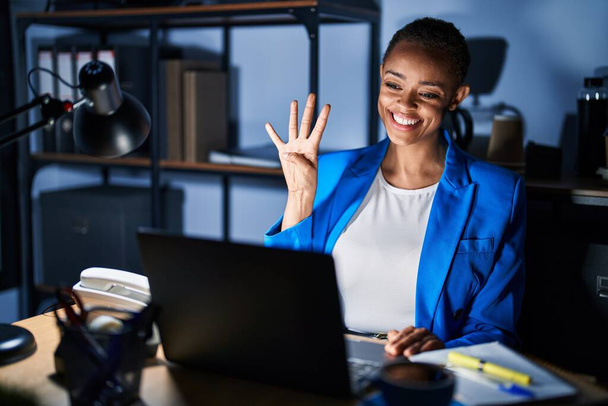 Mulher americana africana bonita trabalhando no escritório à noite mostrando e apontando para cima com os dedos número quatro, sorrindo confiante e feliz.  - Foto, Imagem