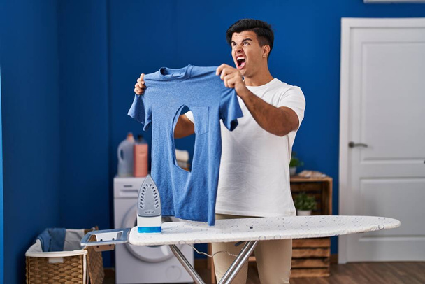 Hombre hispano planchando sosteniendo camisa de hierro quemada en la lavandería enojado y loco gritando frustrado y furioso, gritando con ira mirando hacia arriba.  - Foto, imagen