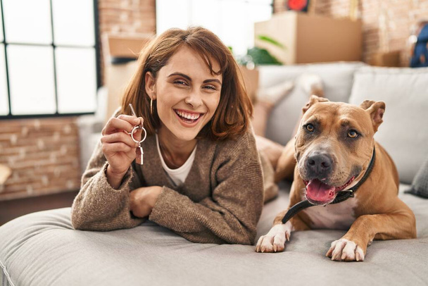 Young caucasian woman holding key of new house lying on sofa with dog at new home - Photo, Image