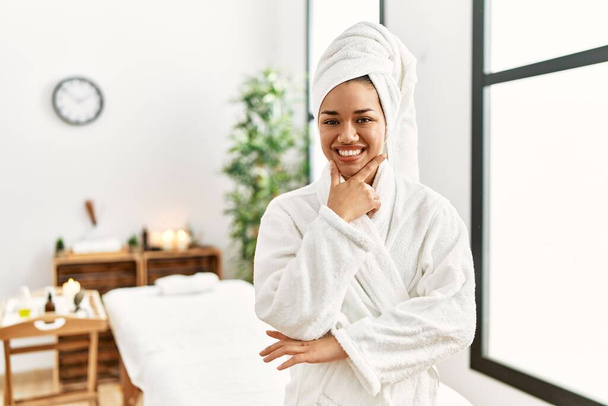 Young brunette woman wearing towel and bathrobe standing at beauty center looking confident at the camera smiling with crossed arms and hand raised on chin. thinking positive.  - Foto, Bild