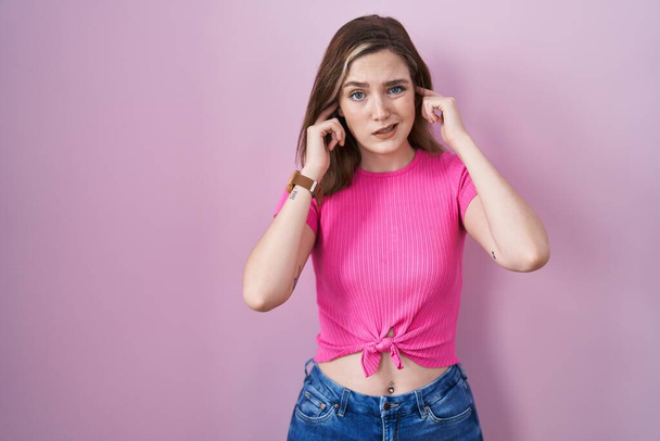 Blonde caucasian woman standing over pink background covering ears with fingers with annoyed expression for the noise of loud music. deaf concept.  - Photo, Image