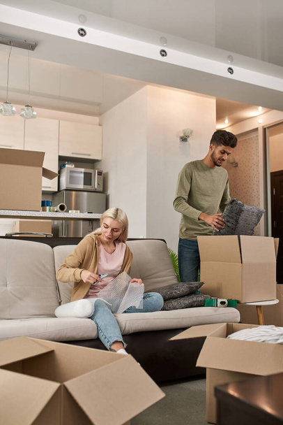 Caucasian girl cut air bubble film with scissors and middle eastern man packing pillows in cardboard box. Young smiling multiethnic couple moving to new apartment. Concept of home relocation - Valokuva, kuva