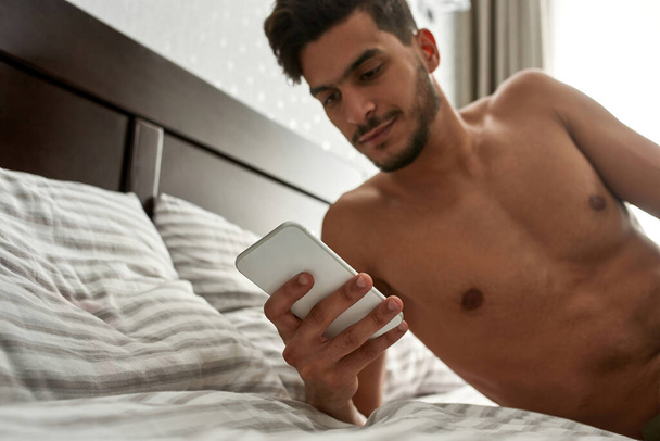 Partial of young concentrated handsome middle eastern man using smartphone on bed at home. Bearded guy with naked torso. Domestic entertainment, rest and leisure. Bedroom in modern apartment. Morning - Valokuva, kuva
