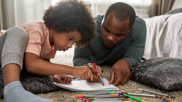Father and son drawing with felt tip pens in sketchbook on carpet with pillows at home. Young black family spending time together. Fatherhood and parenting. Domestic leisure, hobby and entertainment - Photo, Image