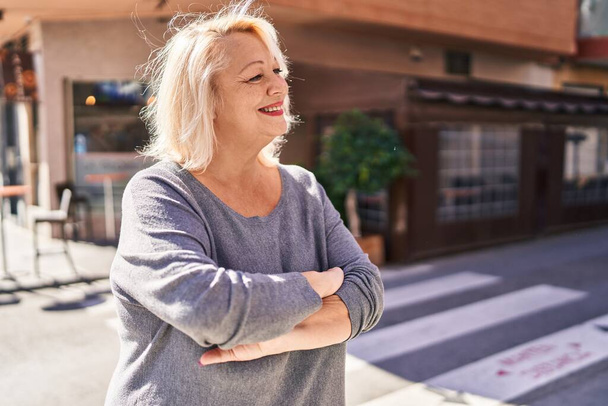 Orta yaşlı sarışın kadın gülümsüyor. Kollarını kavuşturup caddede duruyor. - Fotoğraf, Görsel