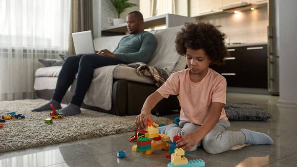 Little boy playing with construction building blocks on floor while his father using laptop on sofa at home. Young black family lifestyle and relationship. Domestic leisure, hobby and entertainment - 写真・画像