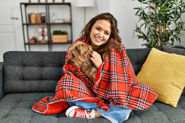 Junge schöne hispanische Frau umarmt Hund, der zu Hause auf dem Sofa sitzt - Foto, Bild