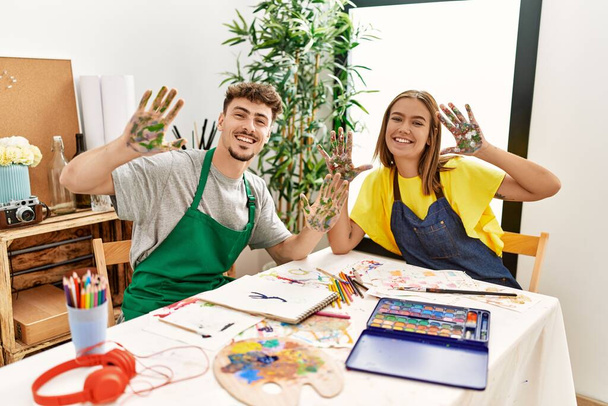 Joven pareja de artistas hispanos sonriendo feliz mostrando las manos pintadas de palma en el estudio de arte. - Foto, Imagen