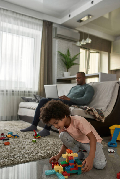 Little boy playing with construction building blocks on floor while his dad typing on laptop on sofa at home. Young black family lifestyle and relationship. Domestic leisure, hobby and entertainment - Foto, Bild