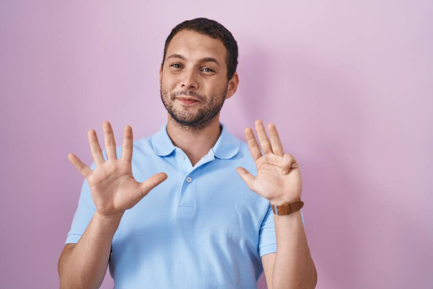 Hispánec stojí nad růžovým pozadím ukazuje a ukazuje prsty číslo devět a usmívá se sebevědomě a šťastně.  - Fotografie, Obrázek