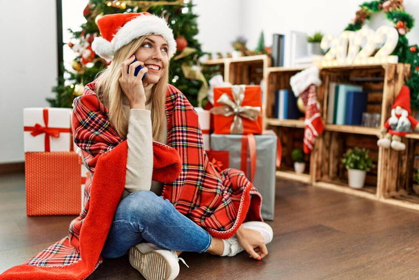 Молодая блондинка разговаривает по смартфону сидя у рождественской елки дома - Фото, изображение