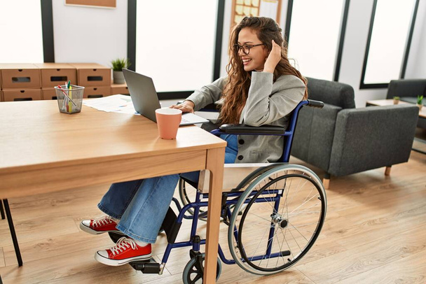 Молодая красивая латиноамериканка бизнес-работница с ноутбуком сидит на инвалидной коляске в офисе - Фото, изображение