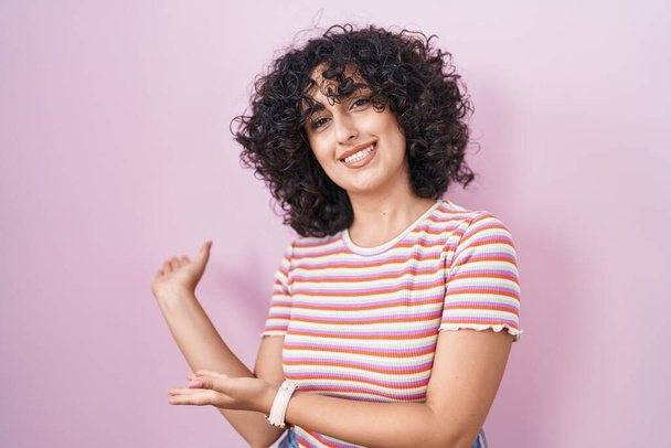 Young middle east woman standing over pink background inviting to enter smiling natural with open hand  - 写真・画像