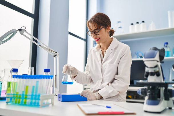 Young caucasian woman wearing scientist uniform measuring liquid at laboratory - Foto, Imagem